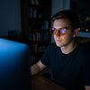 Man working late at a bright iMac in orange-lens blue light glasses, the screen reflected in the lenses