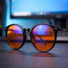 Raven blue light glasses in glossy black TR90 resting on a desk in front of a lit computer monitor