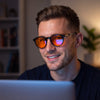 Smiling man at a laptop in a dim room wearing Raven round black blue light glasses with orange lenses