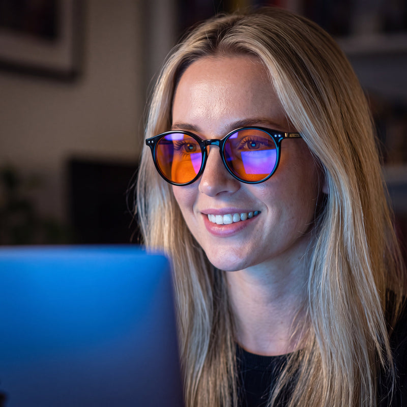 Blonde woman working on a laptop, wearing Raven blue light glasses with black round frames