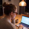 Woman typing at a laptop in the evening wearing the Rowan blue light glasses — glossy black panto TR90 frame